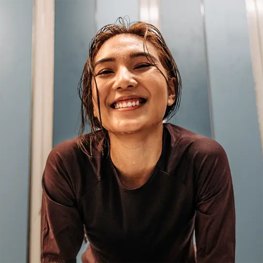 Žena sa vlažnom kosom se smeška. Nosim tamnu sportsku majicu i izgleda sretno, možda nakon fizičke aktivnosti. Pozadina je plava, a osvetljenje naglašava njen vedar izraz lica.