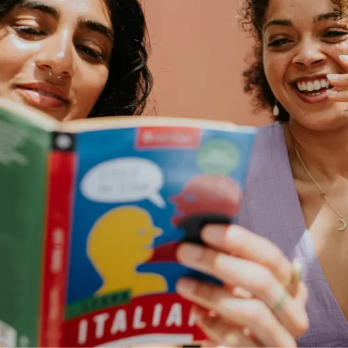 Two women are engaged in conversation while looking at a book titled "Learn Italian." One woman has dark hair and a septum ring, while the other has curly hair and appears to be smiling. The background is a soft, warm color.