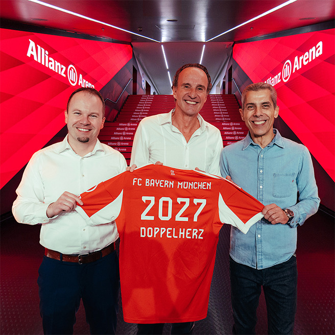 Troje muškaraca stoje zajedno držeći nogometnu majicu s natpisom "FC Bayern München 2027 DOPPELHERZ". Pozadina je stadion Allianz Arena. Svi se osmjehuju, odjeveni su u ležernu odjeću.