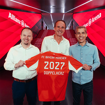 Troje muškaraca stoje zajedno držeći nogometnu majicu s natpisom "FC Bayern München 2027 DOPPELHERZ". Pozadina je stadion Allianz Arena. Svi se osmjehuju, odjeveni su u ležernu odjeću.